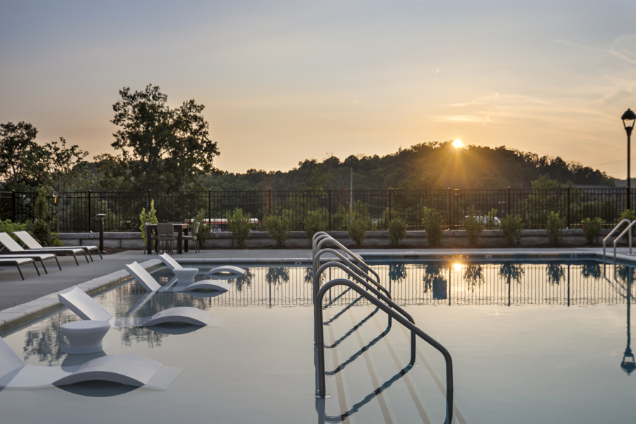 Resort-style apartment pool with in-water loungers, tanning ledge, and scenic sunset views overlooking wooded hills at HARMON West Meade.